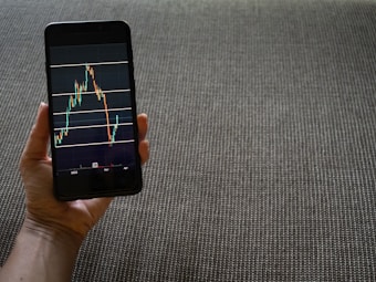 A hand is holding a smartphone that displays a stock market chart with various candlestick patterns and horizontal lines. The phone is held against a textured fabric background, which appears to be a sofa or carpet.