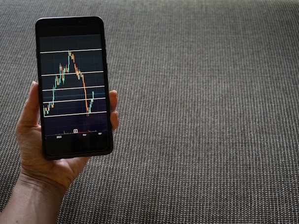A hand is holding a smartphone that displays a stock market chart with various candlestick patterns and horizontal lines. The phone is held against a textured fabric background, which appears to be a sofa or carpet.