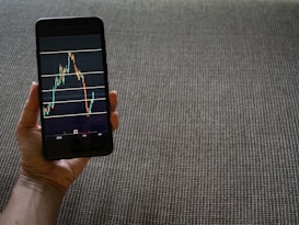 A hand is holding a smartphone that displays a stock market chart with various candlestick patterns and horizontal lines. The phone is held against a textured fabric background, which appears to be a sofa or carpet.