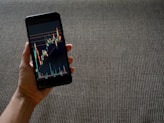 A hand holding a smartphone displaying a colorful line graph with various candlestick patterns, indicative of financial or stock market data. The background consists of a textured, woven fabric surface in neutral tones.