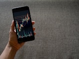 A hand holding a smartphone displaying a colorful line graph with various candlestick patterns, indicative of financial or stock market data. The background consists of a textured, woven fabric surface in neutral tones.