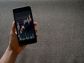A hand holding a smartphone displaying a colorful line graph with various candlestick patterns, indicative of financial or stock market data. The background consists of a textured, woven fabric surface in neutral tones.