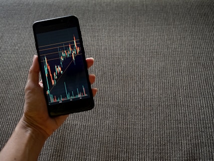 A hand holding a smartphone displaying a colorful line graph with various candlestick patterns, indicative of financial or stock market data. The background consists of a textured, woven fabric surface in neutral tones.
