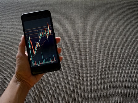 A hand holding a smartphone displaying a colorful line graph with various candlestick patterns, indicative of financial or stock market data. The background consists of a textured, woven fabric surface in neutral tones.