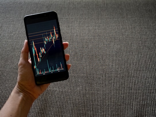 A hand holding a smartphone displaying a colorful line graph with various candlestick patterns, indicative of financial or stock market data. The background consists of a textured, woven fabric surface in neutral tones.