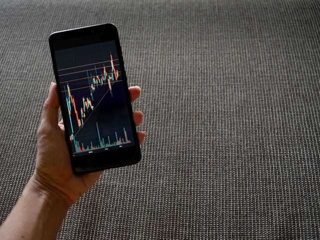A hand holding a smartphone displaying a colorful line graph with various candlestick patterns, indicative of financial or stock market data. The background consists of a textured, woven fabric surface in neutral tones.