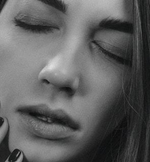 A close-up, black and white photograph of a person with their eyes closed. The facial expression is serene, and the texture of the skin is clearly visible. The image emphasizes the smoothness and subtle highlights on the face, with soft shadows that give a gentle contrast. Long, straight hair frames part of the face, and a hand with neatly manicured nails is gently touching the chin.