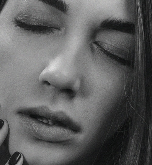 A close-up, black and white photograph of a person with their eyes closed. The facial expression is serene, and the texture of the skin is clearly visible. The image emphasizes the smoothness and subtle highlights on the face, with soft shadows that give a gentle contrast. Long, straight hair frames part of the face, and a hand with neatly manicured nails is gently touching the chin.