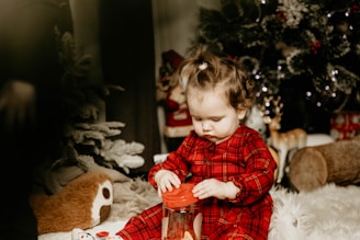 A young child with curly hair, wearing a red plaid outfit, is intently playing with a clear container with a red lid, sitting on a plush white blanket. Surrounding the child, there are festive decorations, including a Christmas tree with lights and ornaments, a plush toy, and a decorative reindeer figure.