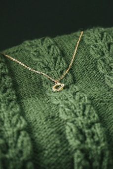 A close-up view of a textured green knitted fabric with a delicate gold necklace resting on top. The necklace features a simple chain and a small, elegant oval pendant.