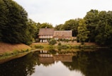 A charming house with a thatched roof is nestled among lush green trees. The building has multiple windows and is situated beside a serene pond that mirrors its reflection. The scene is tranquil with an abundance of greenery surrounding the property.
