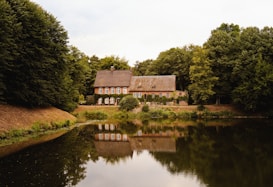 A charming house with a thatched roof is nestled among lush green trees. The building has multiple windows and is situated beside a serene pond that mirrors its reflection. The scene is tranquil with an abundance of greenery surrounding the property.