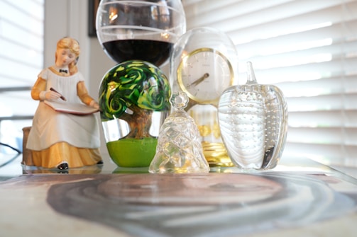 A collection of decorative items is arranged on a glass surface, including a ceramic figurine of a girl writing, a colorful glass object resembling a tree, a glass apple, a clock with a gold rim, and a decorative glass bell. The background consists of partially closed blinds letting in natural light.