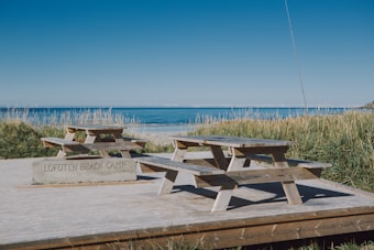 A calm beach setting with two wooden picnic tables sitting on a wooden deck surrounded by tall grasses. In the background, the ocean stretches out under a clear blue sky. A stone with the inscription 'Lofoten Beach Camp' is visible near the tables.