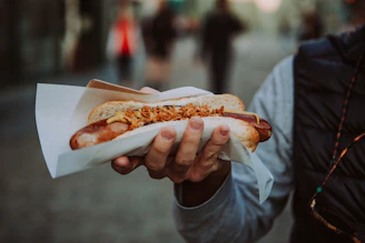 Close-up of a hand holding a freshly grilled Sabrett hotdog topped with mustard and onions against a city backdrop.