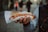 A person holds a hot dog wrapped in paper, topped with mustard and crispy onions. The background is an out-of-focus urban setting.