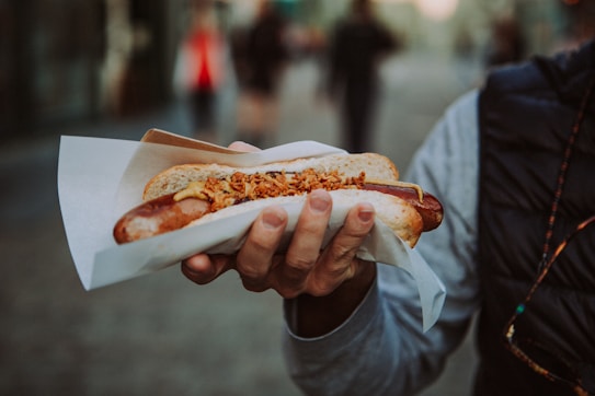 A person holds a hot dog wrapped in paper, topped with mustard and crispy onions. The background is an out-of-focus urban setting.
