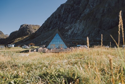 A triangular glass building is situated at the base of rocky mountains, surrounded by a grassy field. Several vehicles and small buildings are nearby, with the structure reflecting the clear blue sky.