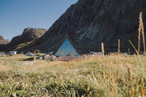 A triangular glass building is situated at the base of rocky mountains, surrounded by a grassy field. Several vehicles and small buildings are nearby, with the structure reflecting the clear blue sky.