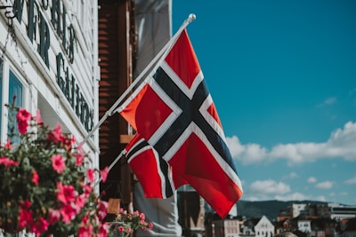 A Norwegian flag waves prominently against a bright blue sky with a few scattered clouds. The flag is mounted on a building facade adorned with vibrant pink flowers in a hanging basket. The surrounding area includes some traditional European architectural elements in the background.