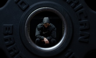 A person wearing a hooded sweatshirt is sitting inside a hollow weight plate. The individual is looking down with a contemplative pose.