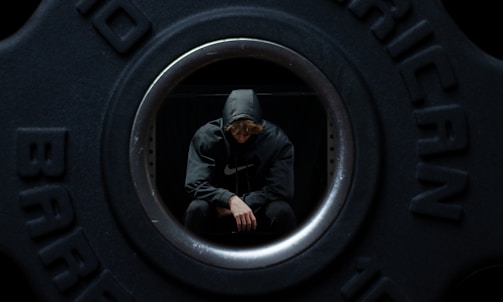 A person wearing a hooded sweatshirt is sitting inside a hollow weight plate. The individual is looking down with a contemplative pose.