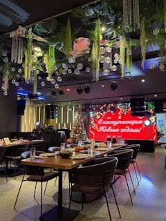 A festive indoor setting featuring a long table set with plates, glasses, and chairs around it. The ceiling is adorned with hanging green plants and ornamental glass balls. In the background, a decorated Christmas tree stands beside a vibrant red sign with text in Cyrillic script, indicating a holiday greeting.
