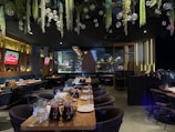 A spacious and elegantly decorated restaurant interior features large tables set with plates and glasses, with greenery and hanging glass orbs from the ceiling. Ambient lighting from wall fixtures and signs create a cozy atmosphere.