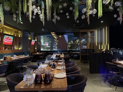 A spacious and elegantly decorated restaurant interior features large tables set with plates and glasses, with greenery and hanging glass orbs from the ceiling. Ambient lighting from wall fixtures and signs create a cozy atmosphere.