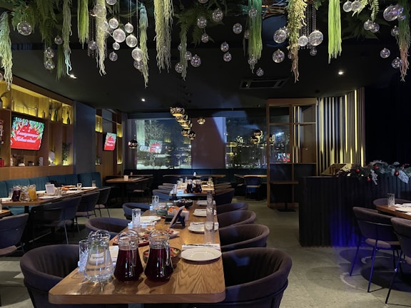A spacious and elegantly decorated restaurant interior features large tables set with plates and glasses, with greenery and hanging glass orbs from the ceiling. Ambient lighting from wall fixtures and signs create a cozy atmosphere.