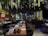 A spacious and elegantly decorated restaurant interior features large tables set with plates and glasses, with greenery and hanging glass orbs from the ceiling. Ambient lighting from wall fixtures and signs create a cozy atmosphere.