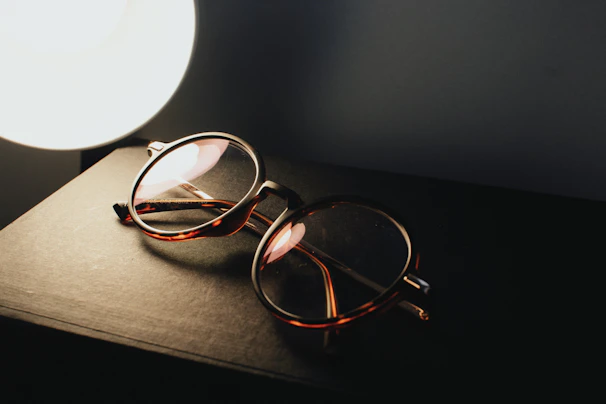 A pair of classic rectangular glasses placed next to a vintage book and reading lamp.