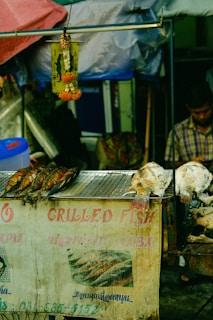 A street food stall displays grilled fish on a metal grill. The stall has a weathered sign with faded text in English and another language, advertising grilled fish. Colorful umbrellas and tarps provide coverage, with a garland hanging from a metal rod above the grill. A person is present in the background, partially visible.