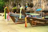 Two traditional Thai long-tail boats are docked at a tropical beach with clear turquoise water. The boats are adorned with colorful ribbons tied to their prows. In the background, there are limestone cliffs and a dense green forest. A couple of people are seen standing near the shore, enjoying the beach.