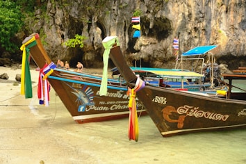 Two traditional Thai long-tail boats are docked at a tropical beach with clear turquoise water. The boats are adorned with colorful ribbons tied to their prows. In the background, there are limestone cliffs and a dense green forest. A couple of people are seen standing near the shore, enjoying the beach.
