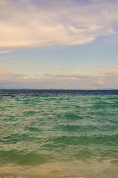A serene seascape with gentle waves in shades of turquoise and deep blue. The sky is partly cloudy with a soft, muted appearance, while the horizon shows faint landforms. A small, yellow buoy floats in the distance, adding a point of interest.