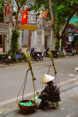 A street scene depicting a person sitting on a small stool, wearing a traditional conical hat. The individual is surrounded by baskets containing goods, possibly for sale. In the background, a decorative gate with flags, including a red flag with a yellow star, is visible. Motorbikes are parked along the street, and trees line the sidewalk.