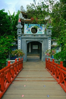 An ornate entrance gate leading to a temple, adorned with intricate carvings and traditional decorations. A red wooden bridge crosses over a water feature, surrounded by lush greenery and flowering plants. The architecture includes stone pillars and colorful motifs, adding to the cultural atmosphere.