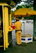 A person in a uniform is standing next to a small kiosk with a large yellow umbrella displaying the logo of VPBank and CSGT. The individual appears to be a traffic officer, wearing a brown uniform with a black belt and dark shoes. The scene is set in an urban environment with a street visible in the background, featuring several moving vehicles and a few pedestrians.