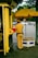 A person in a uniform is standing next to a small kiosk with a large yellow umbrella displaying the logo of VPBank and CSGT. The individual appears to be a traffic officer, wearing a brown uniform with a black belt and dark shoes. The scene is set in an urban environment with a street visible in the background, featuring several moving vehicles and a few pedestrians.