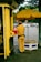 A person in a uniform is standing next to a small kiosk with a large yellow umbrella displaying the logo of VPBank and CSGT. The individual appears to be a traffic officer, wearing a brown uniform with a black belt and dark shoes. The scene is set in an urban environment with a street visible in the background, featuring several moving vehicles and a few pedestrians.