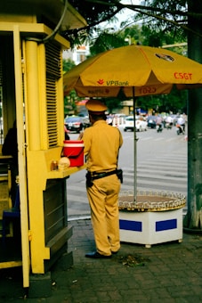 A person in a uniform is standing next to a small kiosk with a large yellow umbrella displaying the logo of VPBank and CSGT. The individual appears to be a traffic officer, wearing a brown uniform with a black belt and dark shoes. The scene is set in an urban environment with a street visible in the background, featuring several moving vehicles and a few pedestrians.