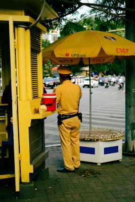 A person in a uniform is standing next to a small kiosk with a large yellow umbrella displaying the logo of VPBank and CSGT. The individual appears to be a traffic officer, wearing a brown uniform with a black belt and dark shoes. The scene is set in an urban environment with a street visible in the background, featuring several moving vehicles and a few pedestrians.