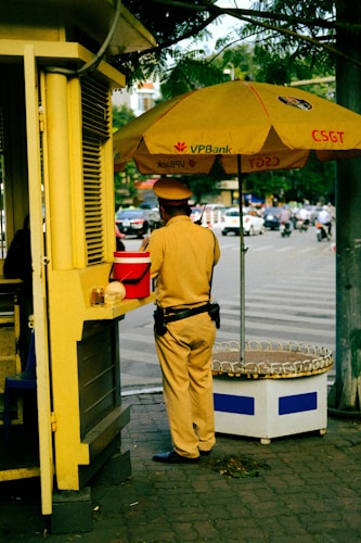 A person in a uniform is standing next to a small kiosk with a large yellow umbrella displaying the logo of VPBank and CSGT. The individual appears to be a traffic officer, wearing a brown uniform with a black belt and dark shoes. The scene is set in an urban environment with a street visible in the background, featuring several moving vehicles and a few pedestrians.