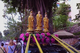 Three golden statues of religious figures are placed on top of a vehicle, decorated with pink flowers and yellow ribbons. The background features a large tree with extensive roots and some greenery. There are also people walking on the left side of the image, and a building is partially visible in the background.