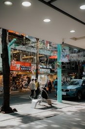 A bustling street scene in an urban area features multiple people walking past shops selling clothing and souvenirs. Vibrant signage with both English and Chinese characters adds to the lively atmosphere. Nearby, a person is sitting on a modern bench under some ambient lighting strung above the street. A blue car is parked to the side, partly in view.