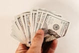 A hand holding a fan of hundred US dollar bills, displaying multiple notes in a splayed arrangement. The bills are crisp and new, showcasing the detailed design and printing.
