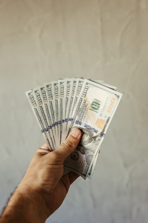 A hand holding a fan of several U.S. hundred-dollar bills, fanned out against a plain background.