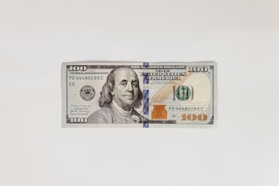 A US one hundred dollar bill featuring an image of Benjamin Franklin on a clean white background. The bill includes typical security features, such as a security strip and a watermark.