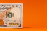 A close-up of a one hundred dollar bill partially placed against a vivid orange background, showing detailed features including the number 100 and parts of the official seal.
